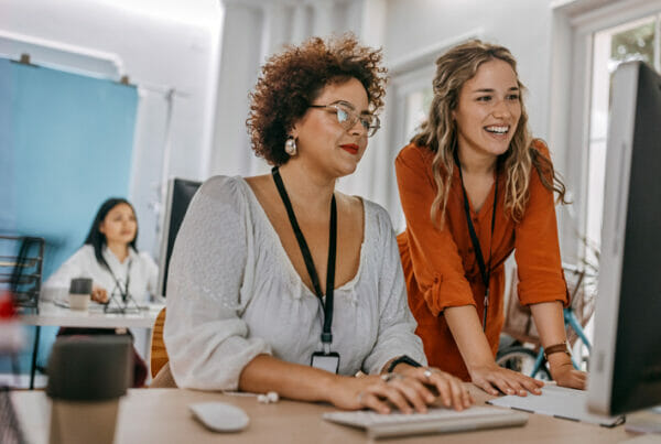 Effective job posting. Two women work together in an office, smiling and looking at the same computer screen.