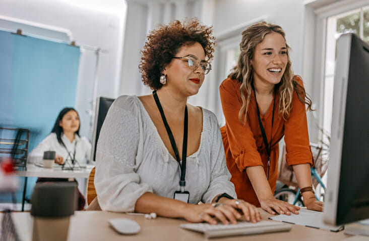 Effective job posting. Two women work together in an office, smiling and looking at the same computer screen.